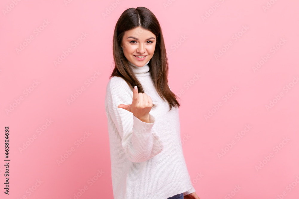 Brunette girl looking playfully, calling with one finger, making beckoning gesture, inviting to come, wearing white casual style sweater. Indoor studio shot isolated on pink background. Brunette girl looking playfully, calling with one finger, making beckoning gesture, inviting to come, wearing white casual style sweater. Indoor studio shot isolated on pink background.