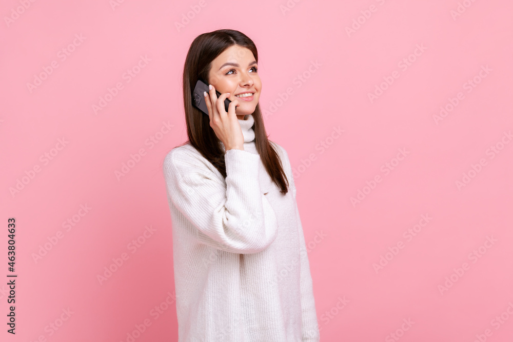 Side view of female talking phone with friend, expressing positive emotions during conversation, wearing white casual style sweater. Indoor studio shot isolated on pink background. Side view of female talking phone with friend, expressing positive emotions during conversation, wearing white casual style sweater. Indoor studio shot isolated on pink background.