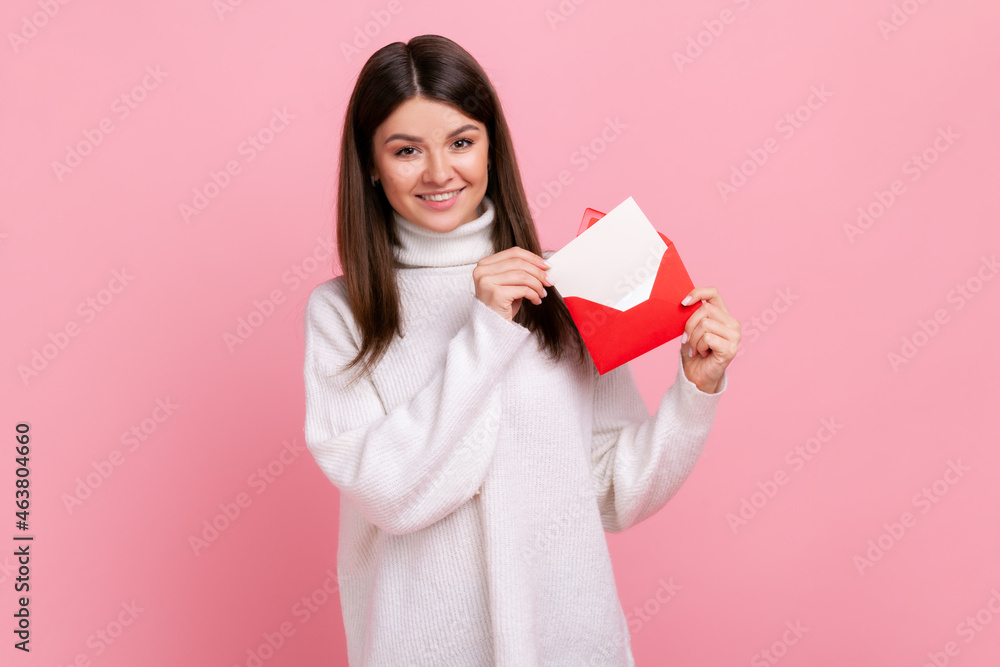 Portrait of woman standing, holding envelope and looking at camera with toothy smile and enjoying, wearing white casual style sweater. Indoor studio shot isolated on pink background. - obrazy, fototapety, plakaty 