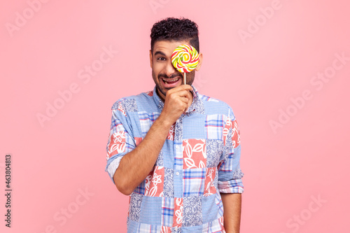 Joyful man with beard wearing blue covering eye with lollipop, having fun, standing with sweet sugary confectionery, looking at camera with toothy smile. Indoor studio shot isolated on pink background