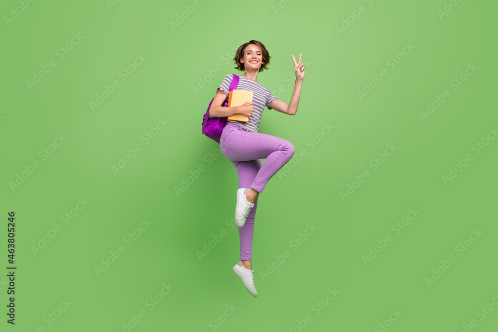 Full body photo of cool young brunette lady hold books jump show v-sign wear bag t-shirt jeans shoes isolated on green background Full body photo of cool young brunette lady hold books jump show v-sign wear bag t-shirt jeans shoes isolated on green background