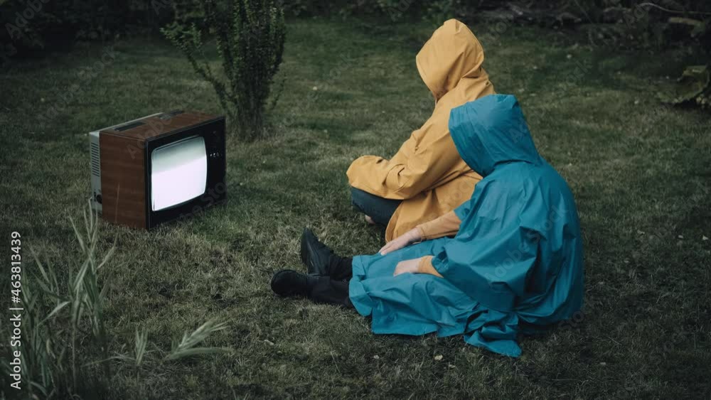 Man and woman in raincoats with hood are sitting on grass and watching an old retro TV. TV is strobing. Concept of post apocalypse, horror, surrealism and mysticism. Zombification, dependence on TV Man and woman in raincoats with hood are sitting on grass and watching an old retro TV. TV is strobing. Concept of post apocalypse, horror, surrealism and mysticism. Zombification, dependence on TV