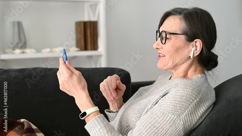 Positive senior mature lady sitting in a relaxed pose on the comfortable couch with a smartphone, mid age woman making a video call, waving hands, greeting friend or family duiring virtual meeting photo