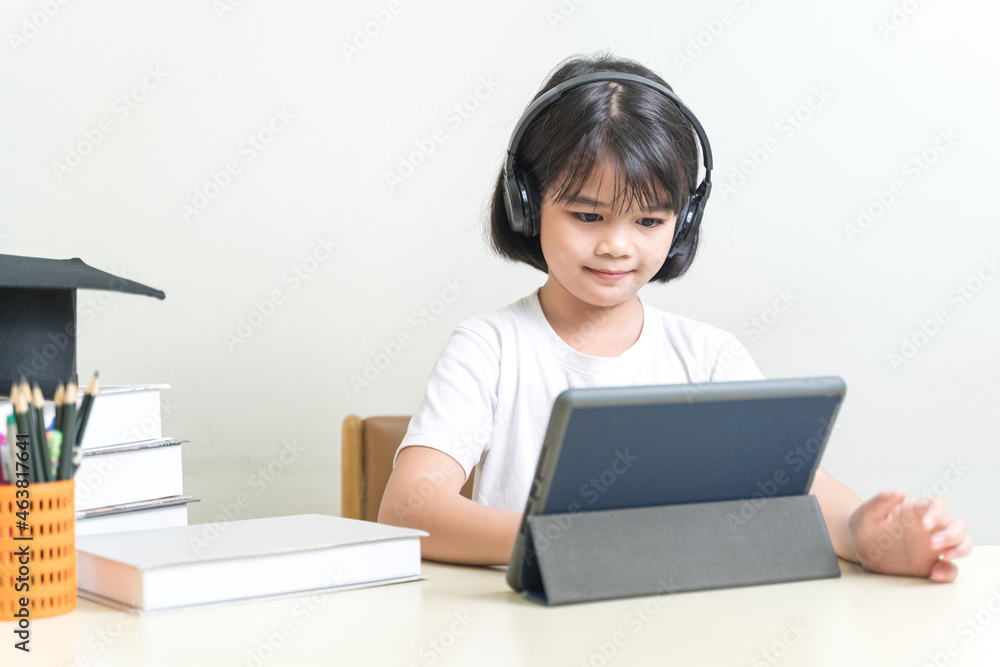 Cheerful Asian little girl student wears headphone, writes on digital tablet to study at home. Education Concept Stock Photo Cheerful Asian little girl student wears headphone, writes on digital tablet to study at home. Education Concept Stock Photo