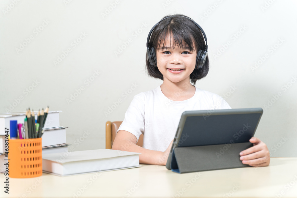 Cheerful Asian little girl student wears headphone, writes on digital tablet to study at home. Education Concept Stock Photo Cheerful Asian little girl student wears headphone, writes on digital tablet to study at home. Education Concept Stock Photo