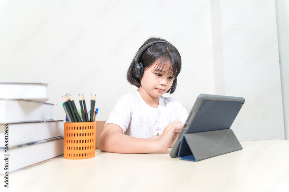 Cheerful Asian little girl student wears headphone, writes on digital tablet to study at home. Education Concept Stock Photo Cheerful Asian little girl student wears headphone, writes on digital tablet to study at home. Education Concept Stock Photo
