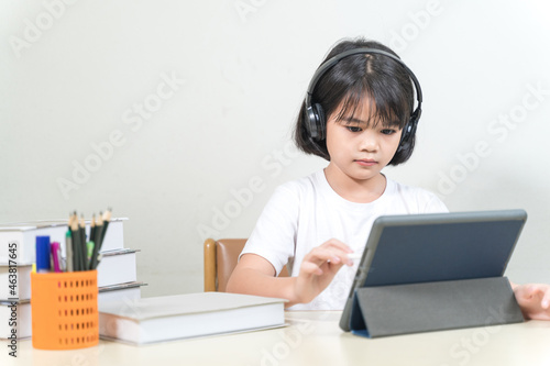Cheerful Asian little girl student wears headphone, writes on digital tablet to study at home. Education Concept Stock Photo