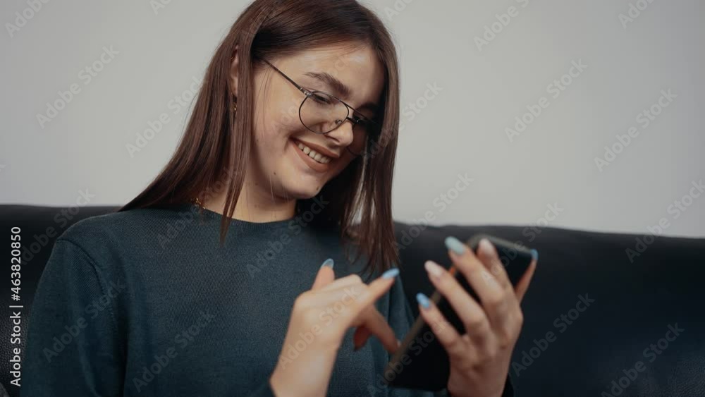 The profile of a freckled young woman, with glasses relaxing at home on the phone, surfing the internet, smiling wide. Dressed in a dark green blouse, sitting on a black sofa. The concept of the woman The profile of a freckled young woman, with glasses relaxing at home on the phone, surfing the internet, smiling wide. Dressed in a dark green blouse, sitting on a black sofa. The concept of the woman