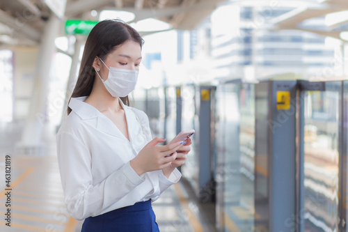 Young Asian business woman who has long hair wears medical face mask protect health from Coronavirus (COVID-19) to prevent disease virus while using the smartphone in her hand to contact the office.