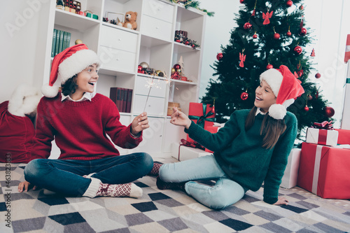 Photo of two funky cheerful kids hold sparkling stick enjoy x-mas atmosphere wear jumper hat in decorated home indoors