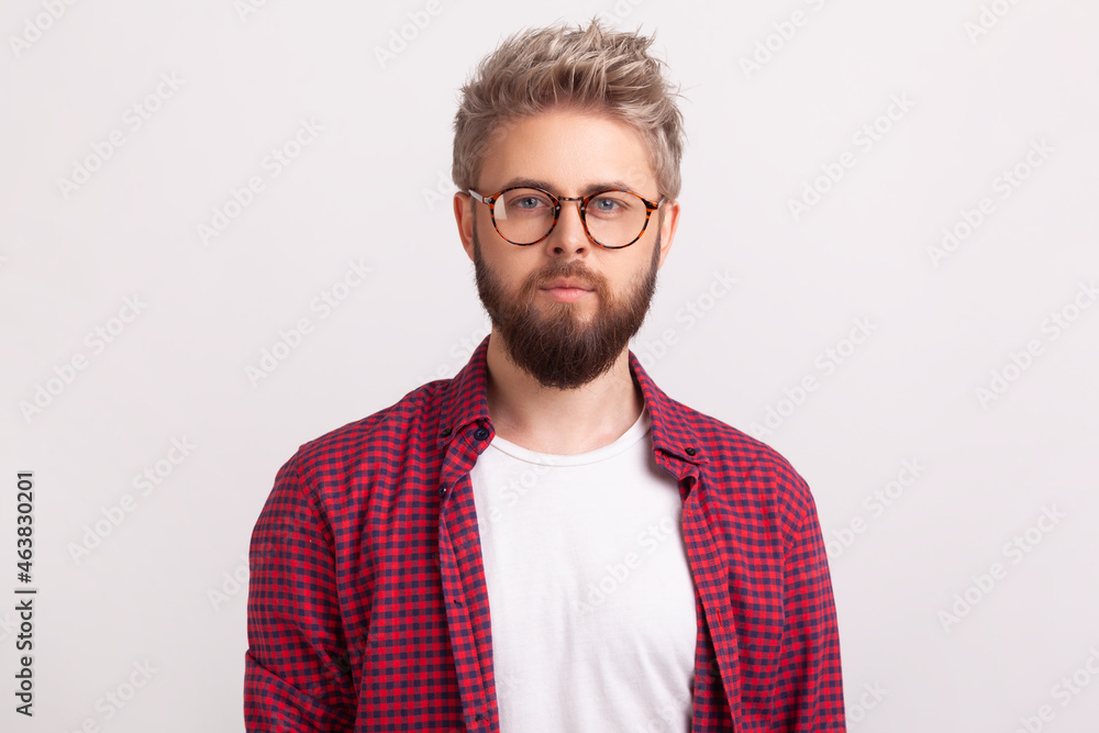 Portrait of handsome self-confident bearded male in eyeglasses wearing red plaid shirt seriously looking at camera. Indoor studio shot isolated on gray background Portrait of handsome self-confident bearded male in eyeglasses wearing red plaid shirt seriously looking at camera. Indoor studio shot isolated on gray background