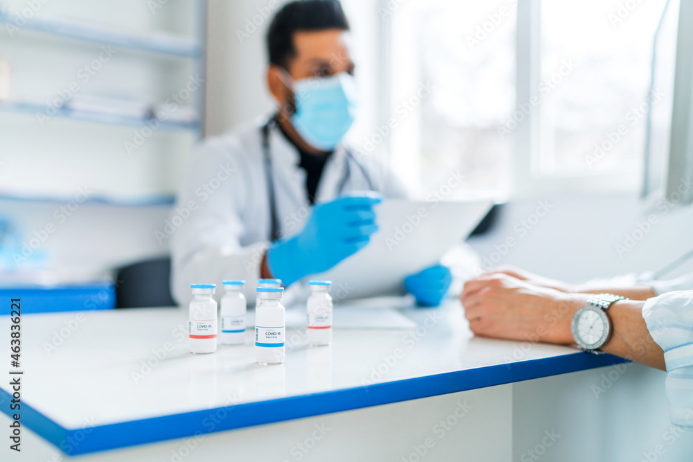 In the focus of the jar with the coronavirus vaccine, on the doctor's desktop vaccine, the doctor consults his patient