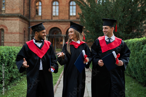 Three smiling gradutes international friends in graduation gowns walking in campus photo