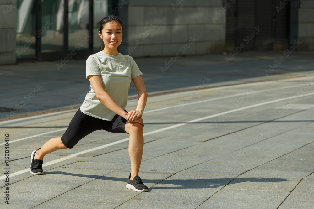 Positive strong Asian woman in tracksuit and shoes does forward dynamic lunges training on sunny city square on summer day Positive strong Asian woman in tracksuit and shoes does forward dynamic lunges training on sunny city square on summer day