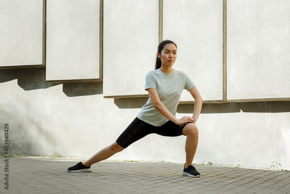 Concentrated Asian lady in tracksuit and sneakers does side dynamic lunges training on pavement near concrete wall in street Concentrated Asian lady in tracksuit and sneakers does side dynamic lunges training on pavement near concrete wall in street