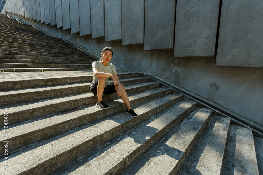 Attractive Asian woman in tracksuit and sneakers sits resting on empty grey steps casting shadow after outdoor training in sunny city
