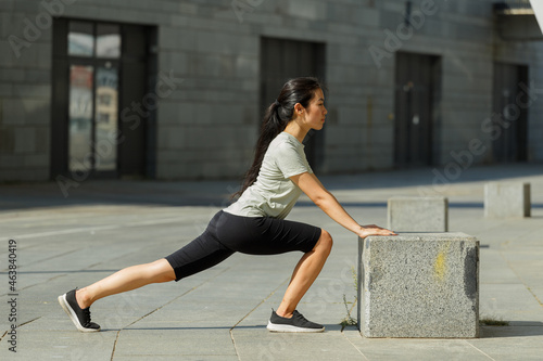 Strong Asian woman with long hair in tracksuit does forward dynamic lunges leaning on stone block on city street side view