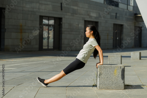 Stylish Asian woman with long ponytail in sports clothes does reverse press-ups leaning on grey stone block on sunny street