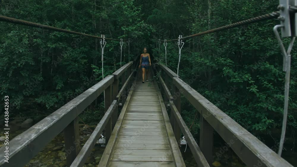 Woman Walking Along Suspension Bridge Alone in Picturesque Green Forest Setting. Slow Motion of Dark Haired Woman Walk on Suspension Bridge in Thick Green Forest. No Other People Are in The Shot. Woman Walking Along Suspension Bridge Alone in Picturesque Green Forest Setting. Slow Motion of Dark Haired Woman Walk on Suspension Bridge in Thick Green Forest. No Other People Are in The Shot.