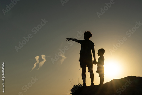 Mother and son against the light silhouetted by the sun pointing with their arms towards the horizon at sunset photo