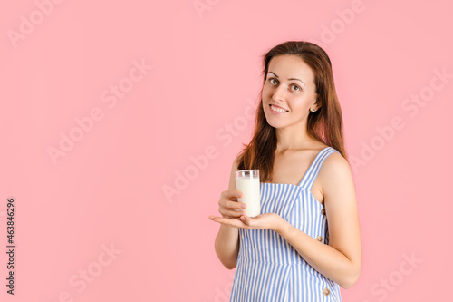 A smiling girl in a blue dress holds a glass of milk in her hands standing on a pink background