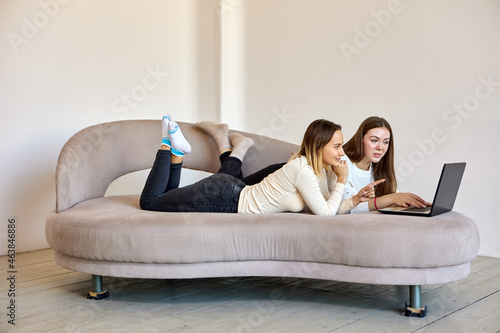 Two women look at laptop during lie on divan in lounge.