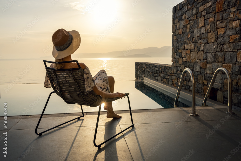 relaxing vacation - woman sitting in chair on terrace in front of pool and enjoying sunrise at luxury villa - obrazy, fototapety, plakaty 