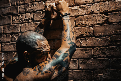 Photography A bald gang member in tattoos stands facing the wall with his hands up in handcuffs