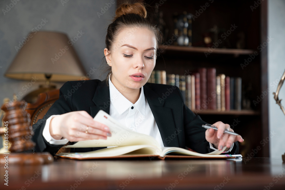 Young female lawyer during work in chamber. Gavel and Themis statue  on the brown shining desk.