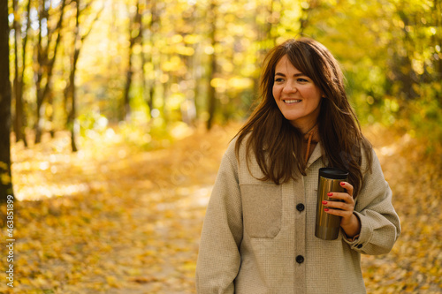 A young woman in a long shirt with a thermo mug in her hands walks through the autumn forest. Natural landscape. Beautiful forest. Autumn day.