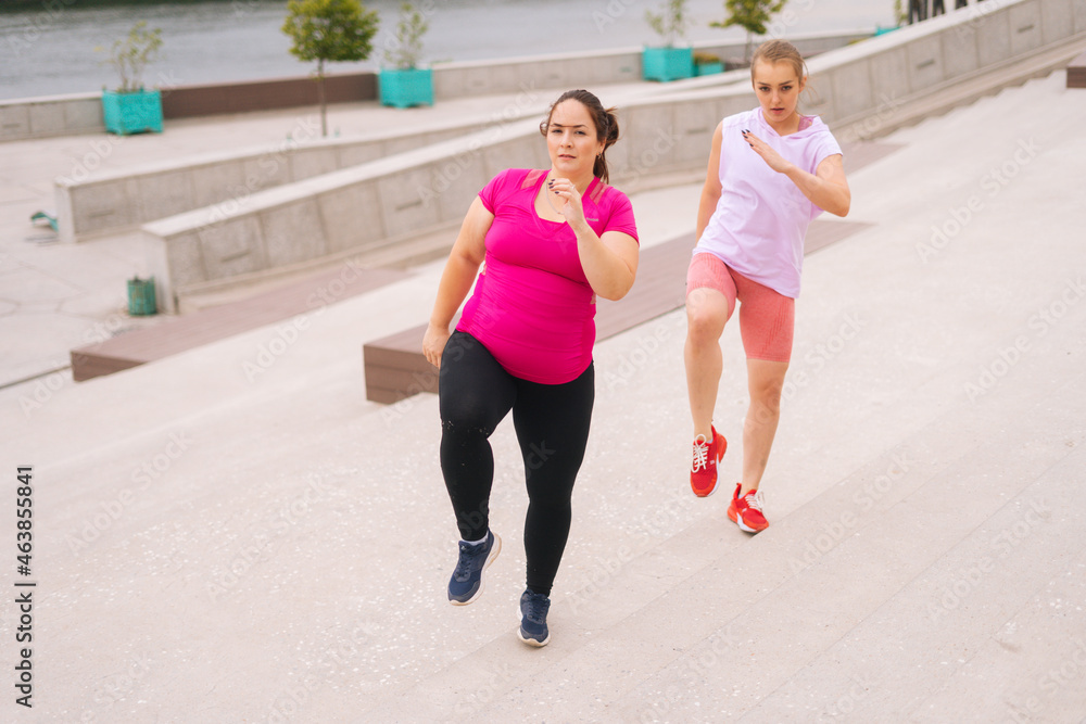 Personal fitness female trainer helping fat woman lose weight outside running on city stairs in summer morning. Instructor giving training to overweight young woman in cloudy summer day.
