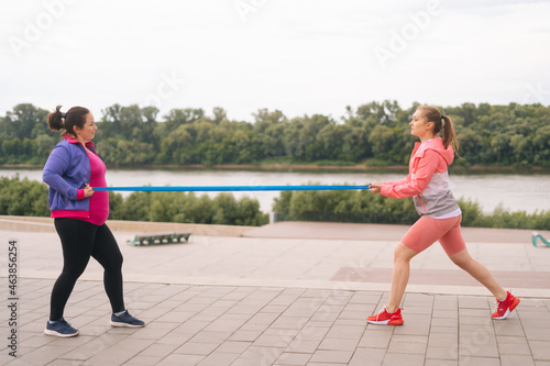 Wide shot of fat young woman doing squats exercises using resistance band for weight loss with personal trainer outdoor in cloudy summer evening. Instructor help overweight woman lose weight.