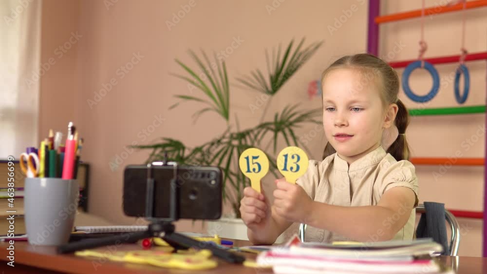 A preschool girl shows plates with numbers, looks into a smartphone.She answers the question. A preschool girl shows plates with numbers, looks into a smartphone.She answers the question.