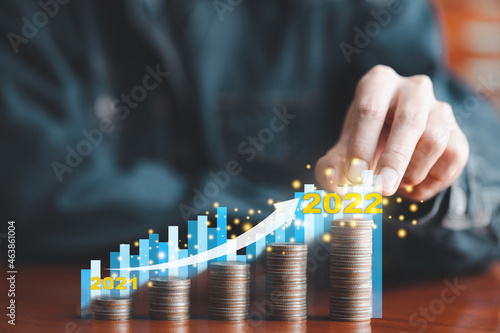 Businessman putting coin on stack coins with blue bar graph virtual image. Analysis of business growth trends previous year 2021 to current year 2022 concept. Save money, Start investing for future