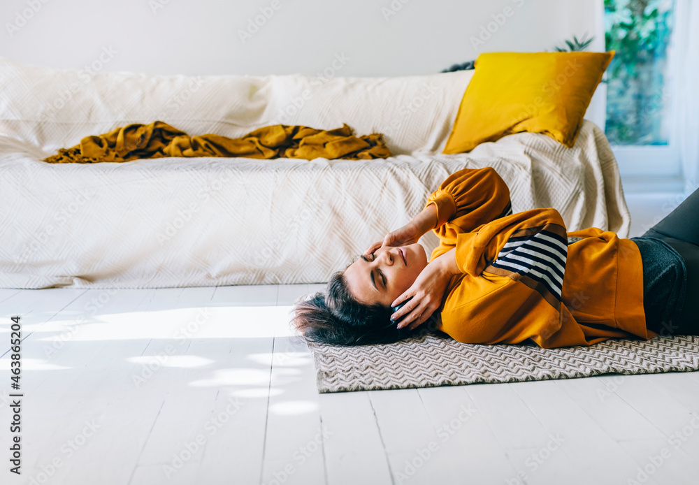Woman in casual clothing with headphones lying down the floor in her living room and enjoys listening the music. Leisure time of single woman relaxing in isolation streaming songs from player. - obrazy, fototapety, plakaty 