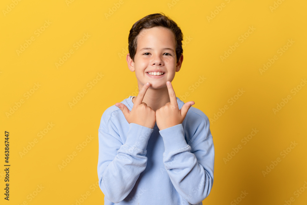 Little boy smiles, pointing fingers at mouth isolated over yellow background. Little boy smiles, pointing fingers at mouth isolated over yellow background.