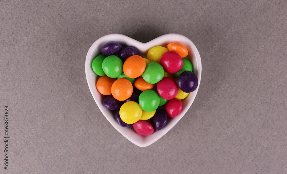 Multicolored round candies in a white plate in the shape of a heart on a gray background Multicolored round candies in a white plate in the shape of a heart on a gray background