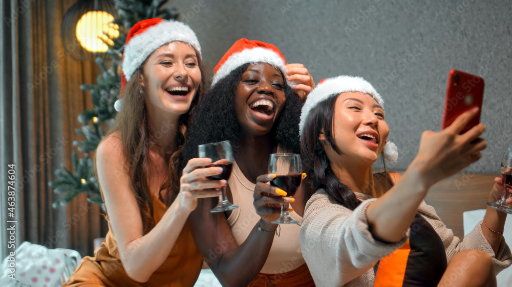 Obraz premium young girls European, Asian and African in red New Year's clothes and hats sit on a bed at a Christmas tree holding glasses of wine made photo on a phone, smiling and celebrating Christmas