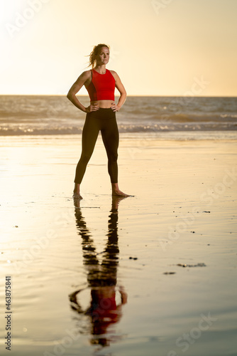 Attractive Strong Young Fit Athletic Blonde Woman in Yoga Pants Confident Pose Reflection in Water Wearing Red Shirt Yoga Pants on beach at Sunset with Yellow Sky Background