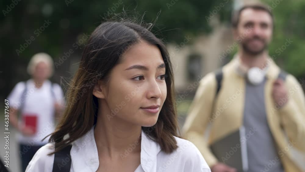 Close-up of smiling young woman walking through campus among multiracial classmates blurred on background. Group of multi-ethnic high school students walking along park alley before study Close-up of smiling young woman walking through campus among multiracial classmates blurred on background. Group of multi-ethnic high school students walking along park alley before study