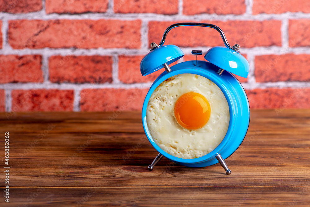 Close-up photo of blue alarm clock with fried egg inside is on wooden table and brick wall background. Wake up and breakfast concept