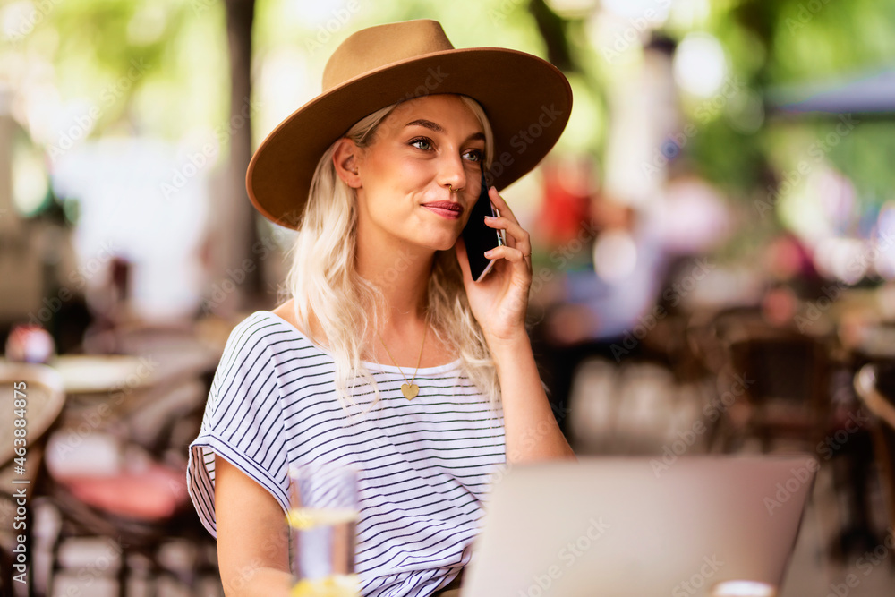 Blond haired woman wearing hat and making a call while sitting at outdoor cafe