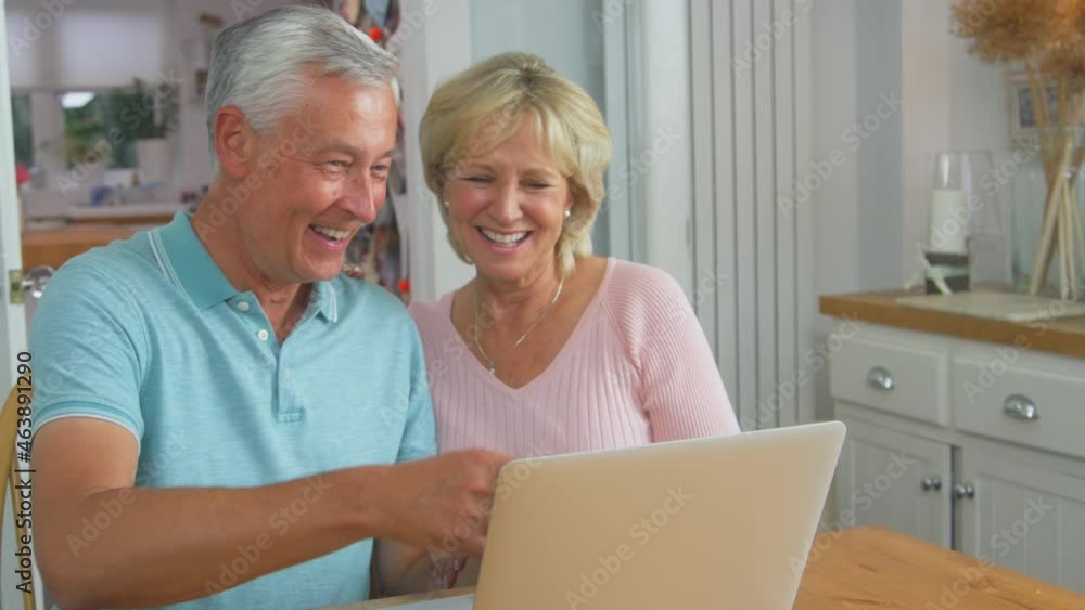 Smiling senior retired couple sitting around table at home making video call on laptop and waving to family- shot in slow motion Smiling senior retired couple sitting around table at home making video call on laptop and waving to family- shot in slow motion