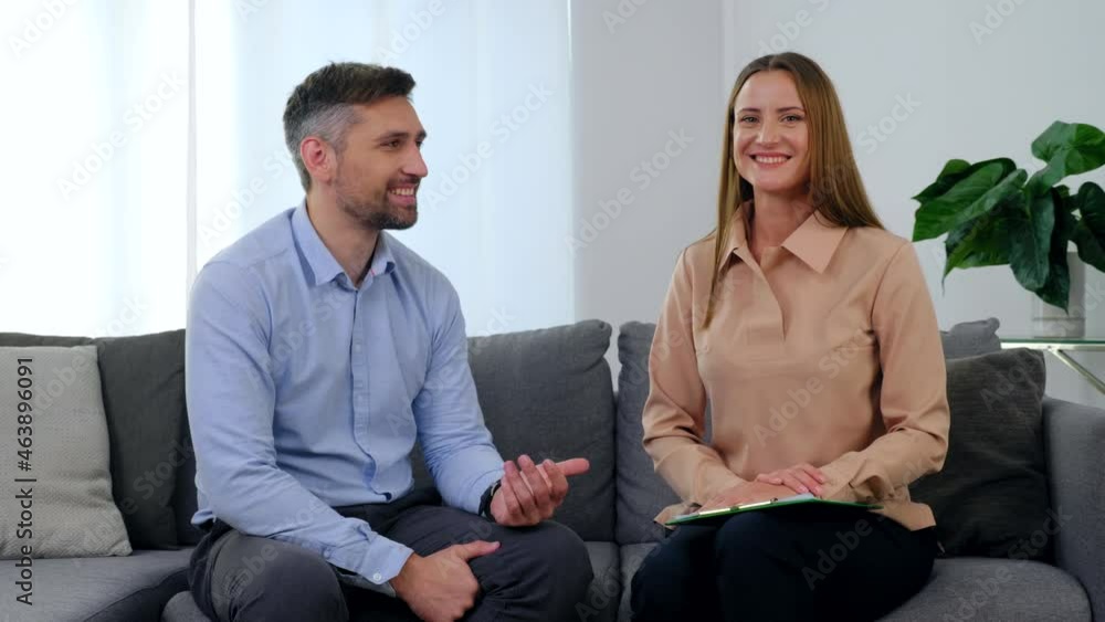 Man patient and woman psychologist sitting on couch talking and turn to camera with smile after having passed positive psychotherapy session in clinic office. Mental health problems after covid-19 Man patient and woman psychologist sitting on couch talking and turn to camera with smile after having passed positive psychotherapy session in clinic office. Mental health problems after covid-19