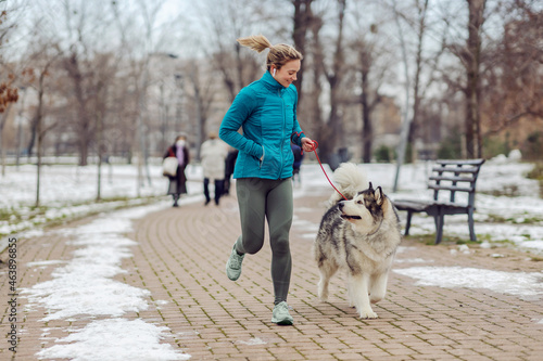 Sportswoman jogging together with her dog in park on snowy weather. Winter fitness, pets, friendship