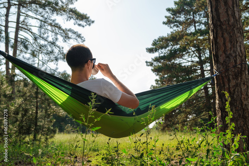A tourist in a hammock is resting in the forest and drinking from a cup.