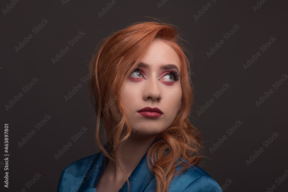 Young beautiful woman with red hair looks up with a blue jacket on a gray background. Young redhead woman smiles with autumn colors Young beautiful woman with red hair looks up with a blue jacket on a gray background. Young redhead woman smiles with autumn colors