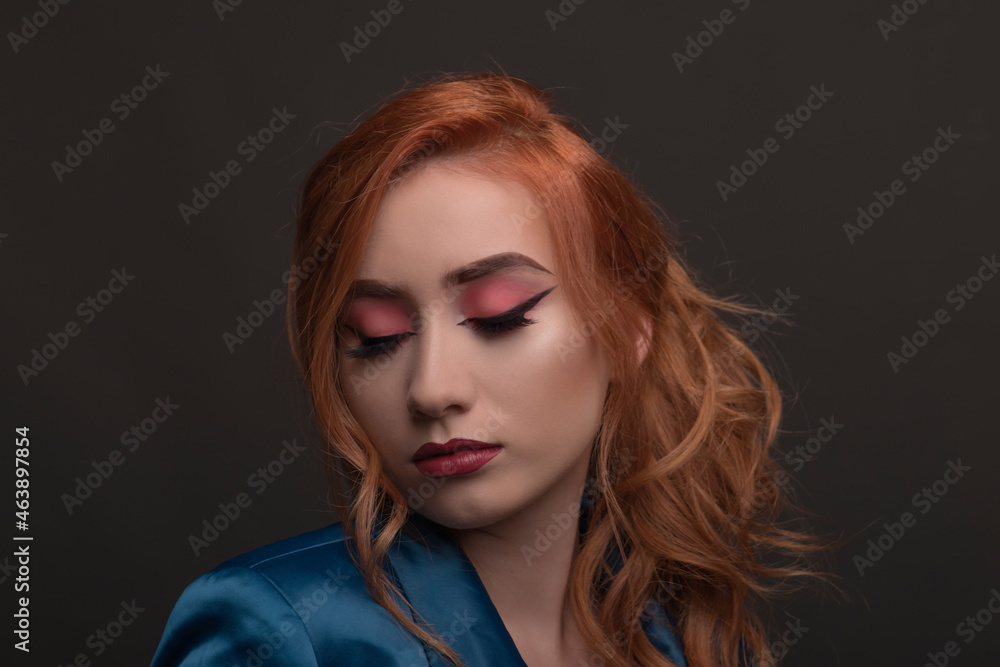 Fototapeta premium Young beautiful woman with red hair looks down with a blue jacket on a gray background. Young redhead woman smiles with autumn colors