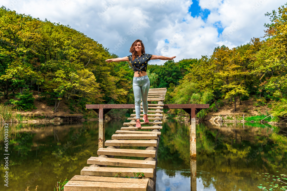 Naklejka premium A young woman on a wooden ladder bridge on a forest mountain lake. Amazing summer landscape with a lake and a panoramic view of a mountain lake