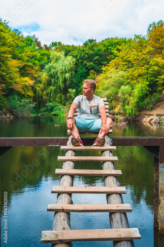 A young man on a wooden ladder bridge on a forest mountain lake. Amazing summer landscape with a lake and a panoramic view of a mountain lake © KseniaJoyg